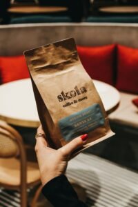 A hand holding a Skola coffee pack with a café setting in the background.